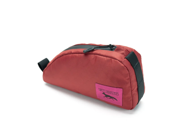 A Swift Industries Moxie Top Tube Bag is shown in a reddish orange material with a black zipper and a pink logo